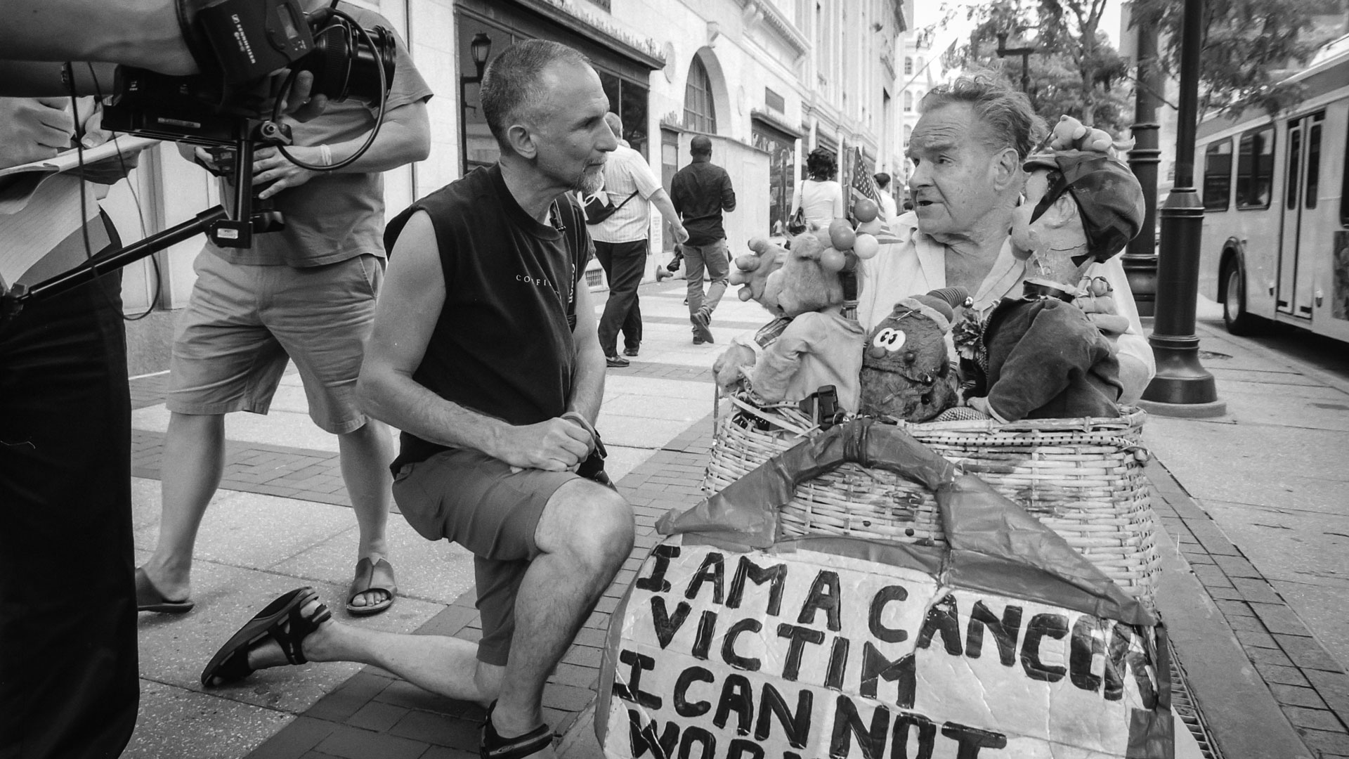 Willie Baronet kneeling to talk with a man on the street beside his handmade sign reading I am a cancer victim I cannot work, while a film crew records